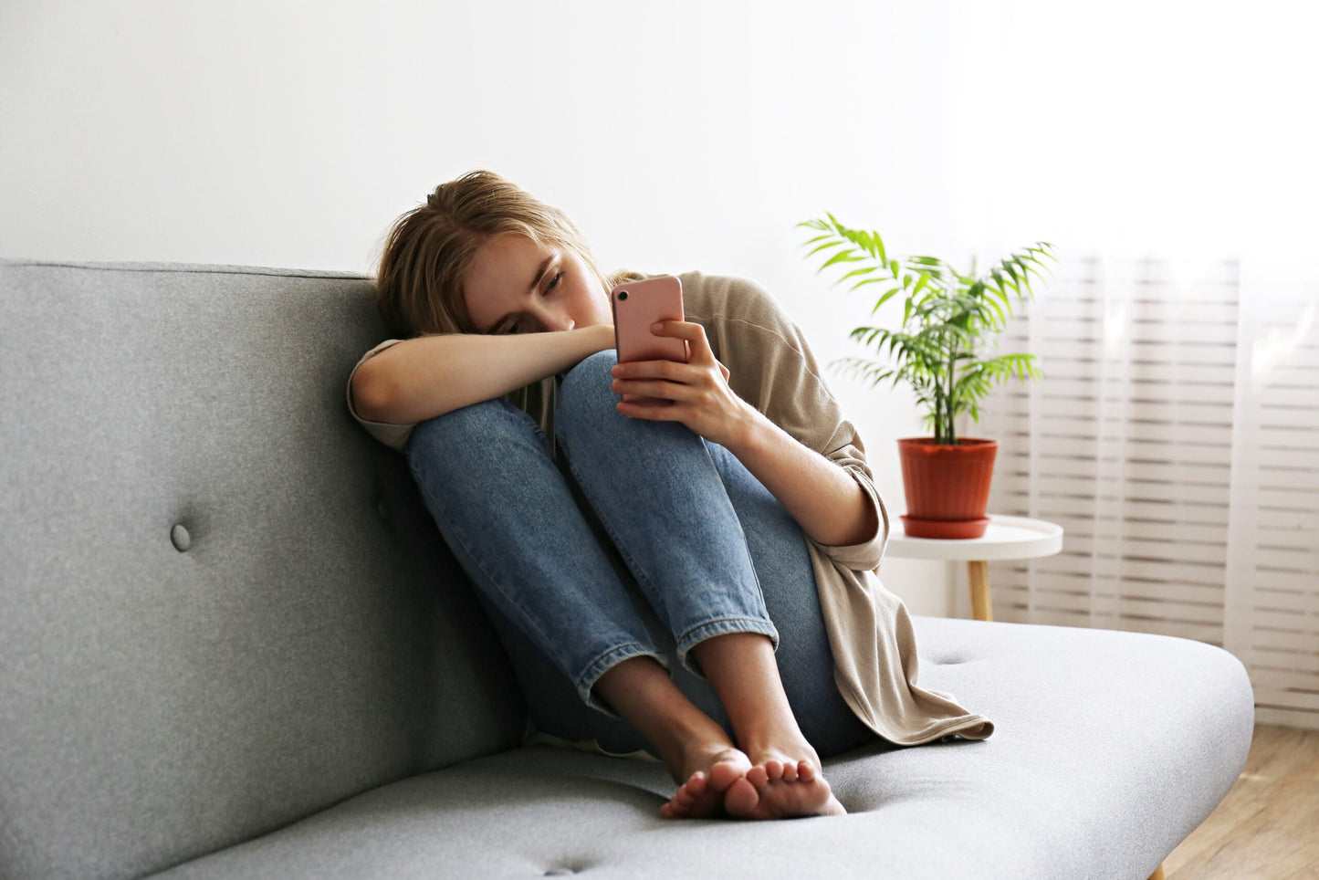 Photo of a depressed looking young woman sitting on a couch looking at her cell phone